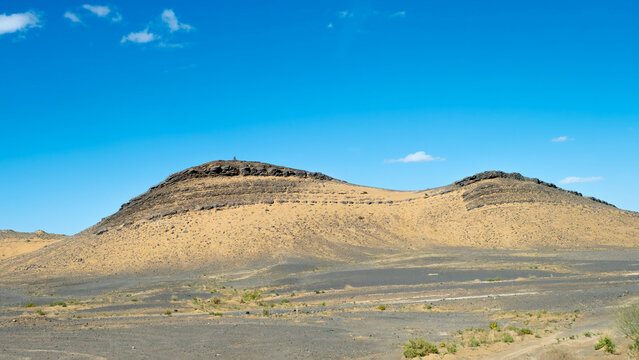wide angle view of dark volcanic rock hills and a stony desert plain under a bright blue sky with sparse clouds in the Sahara region of Morocco