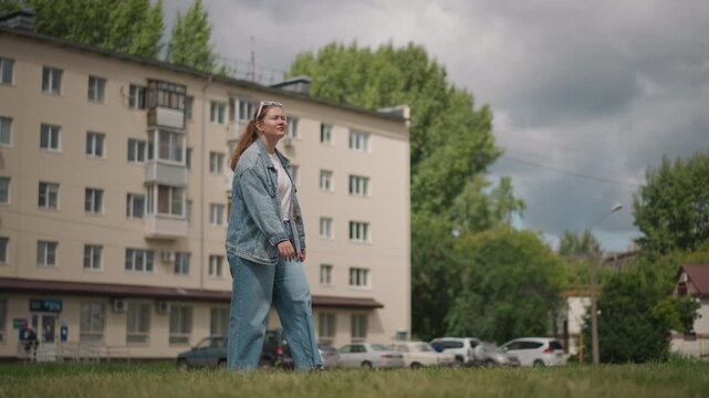 Young woman walking by apartment building, slow stroll across grassy verge in denim outfit, lowangle wide shot under overcast sky, parked cars and trees in background, frames portray student, artist