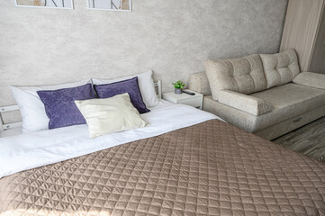 Naklejka premium cozy bedroom with a double bed, quilted beige bedspread, purple cream pillows, beige sofa, white bedside table with plant, textured walls, framed art, neutral tones