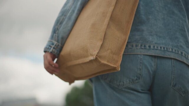 White woman walking with woven tote in denim jacket and jeans under overcast sky, slow paced market stroll, closeup of hand gripping straw bag, textured weave and earthy tones, sustainable reusable