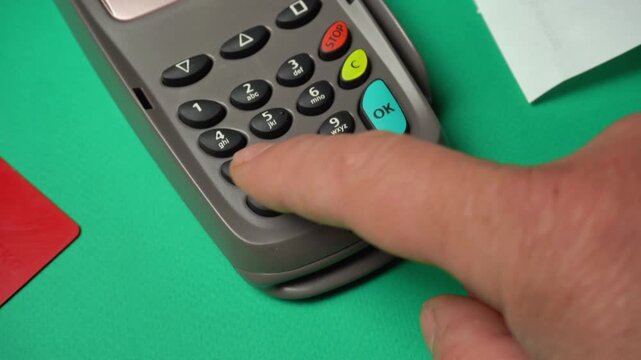 Hand pressing keys on a payment terminal with green background and visible credit cards and receipts nearby during a transaction process