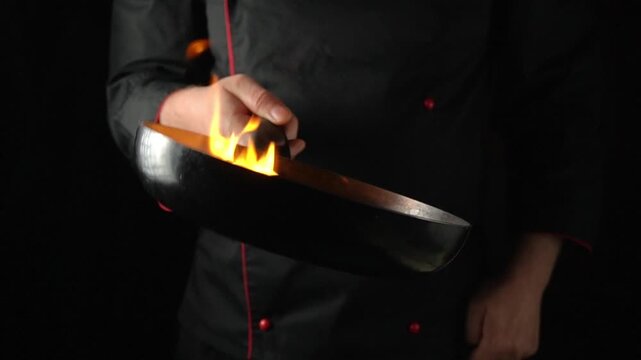 Chef in black uniform performs flambe technique with a frying pan, showcasing flames and colorful vegetables in a dark kitchen setting
