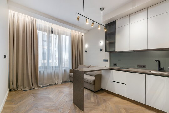 A cozy living area connects to a kitchen space. Large curtains frame the window. Simple furniture is arranged for comfort. Soft lighting hangs from the ceiling in a city apartment