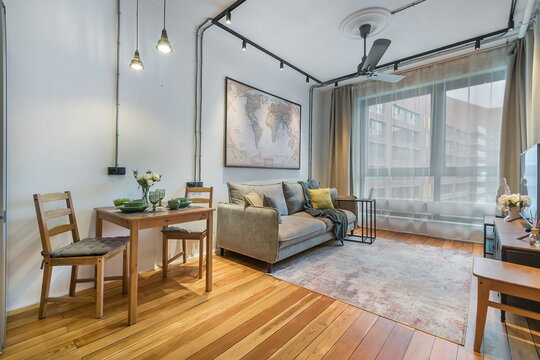 A living room features a sofa and a dining table with chairs. A large window shows city buildings outside. Warm light hangs from the ceiling