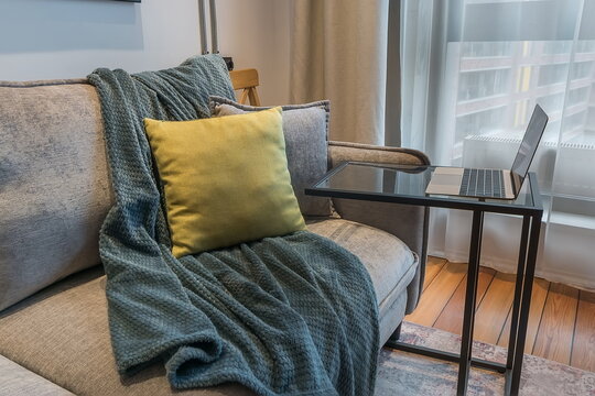 A couch is arranged in a living room. A green blanket drapes over it with a yellow pillow placed on the side. A laptop rests on a small table next to the couch