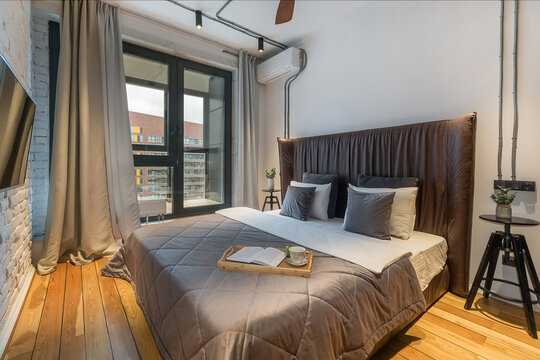 A modern bedroom features a bed with a brown quilt and pillows. A table holds a book and cup. There is a large window with curtains letting in sunlight and wooden floors