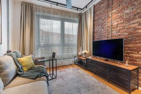 Couch sits on hardwood floor in a living room. A small table stands beside the couch. A television is on a shelf. Large windows show buildings outside. Curtains are drawn