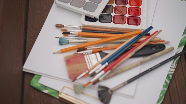 Brushes and palette on wooden table. Watercolor artistry setup with sunlight illuminating materials. Painting essentials carefully placed on rustic surface under warm beams of sunlight