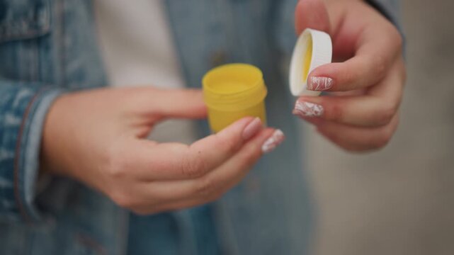 Outdoor hands unscrewing yellow jar young woman in denim jacket with floral nails removing inner seal, preparing balm or ointment, overcast background, practical onthego care, pocket routine
