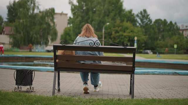 White woman sitting on bench facing fountain, denim jacket, small bag and luggage on ground, trees and water background, contemplative posture, poetic quiet, gentle overcast lighting