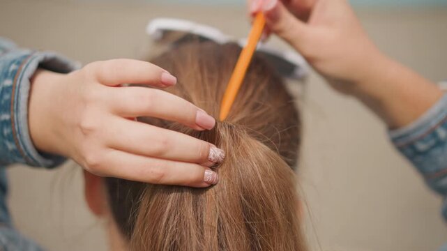 Caucasian artist securing ponytail with pencil closeup, hands inserting orange pencil through brown hair elastic, manicure visible, textured strands, improvisational styling in urban setting