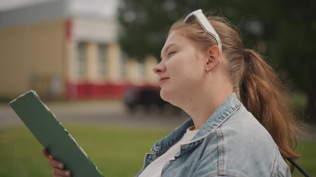 White woman near campus holding folder, denim jacket, green binder clasped to chest, eyes closed with satisfied smile, ponytail and headband fluttering in breeze, candid profile shot conveying relief
