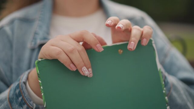 Woman with vibrant nail art. Lady with glittery manicure studying sketches on park bench. Young woman in denim jacket browsing through colorful sketchbook outdoors in peaceful park setting