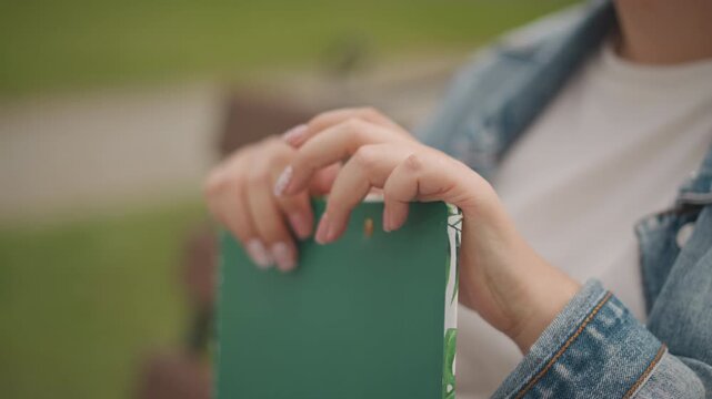 Outdoor hands gripping green notebook on bench, denim jacket sleeve, floral acrylic nails, soft sunlight and grass bokeh, casual waiting moment, reflective mood, candid leisure scene, journal focus