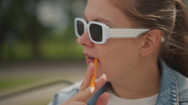 Caucasian woman in sunglasses tasting candy stick, denim jacket profile by park pathway, closeup of lips and orange candy, playful snacking moment, soft bokeh trees, relaxed streetstyle aesthetic