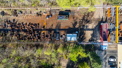 Naklejka premium Top down aerial view of an industrial scrap yard with metal containers, overhead gantry crane, blue truck with red trailer, and parking area surrounded by green trees in bright sunlight.