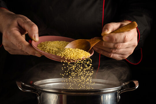 Hands are seen pouring millet from a plate into a pot. Steam rises from the pot as food is being prepared. The scene is focused on the activity of cooking