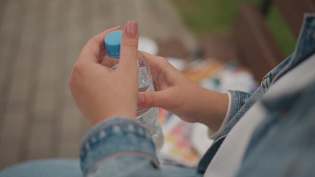 White woman opening plastic water bottle, closeup of hands twisting blue cap, denim sleeves and manicured nails, picnic items blurred on bench, candid hydration moment, summer sunlight