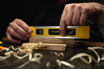 A person focuses on leveling a piece of wood using a spirit level tool. Small wood shavings are scattered on the workbench. The scene shows a workshop with dim lighting © Віталій Б.