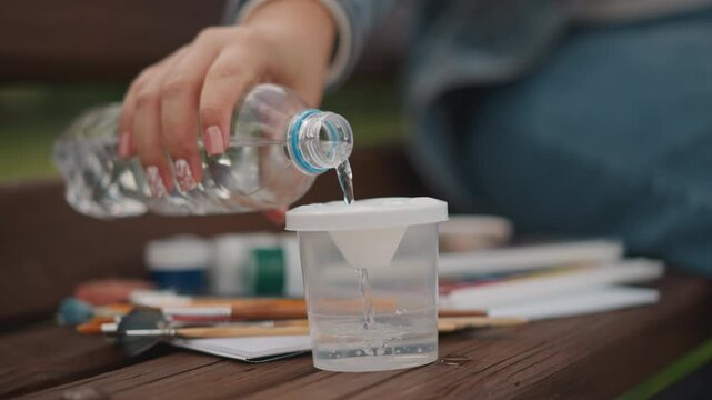 Hands pouring water into cup plastic bottle tipping disposable cup filling brushes and paint tubes on wooden bench denim jeans in background soft sunlight tranquil park preparing watercolor session