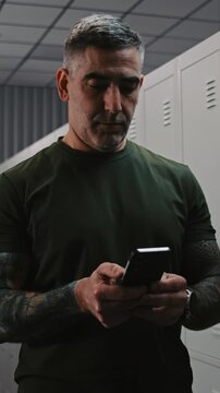 Athletic Military Man in Camouflage Cap and Tactical Gear Uses Smartphone to Send Important Updates or Contact Relatives at Army Base Locker Room Area. Concept of Modern Warfare Communication