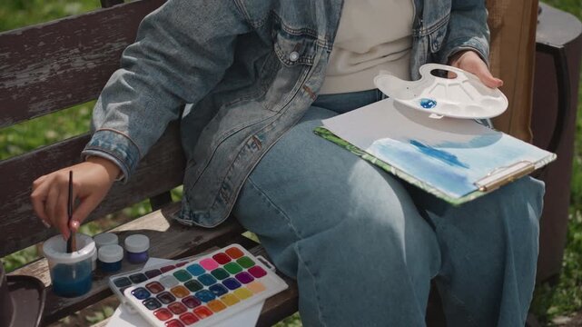 White woman painting on park bench mixing blue hues on plastic palette dipping brush into water cup sketchbook on lap denim jacket and jeans sunlight through leaves relaxed focused mood outdoor