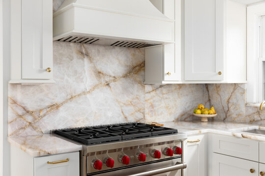 A white kitchen range hood detail with a marble countertop and backsplash, stainless steel oven, and gold accents.