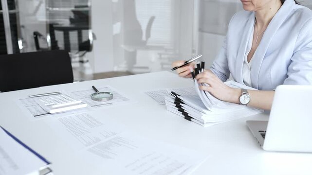 Woman's hands signing and checking financial documents, surrounded by a high stack of paperwork, a calculator, and a laptop, symbolizing busy office work, auditing, and bureaucracy. Audit and taxes