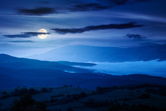 foggy carpathian countryside scenery in summer at night. mysterious view of mountainous landscape with rural fields on rolling hills. full moon over fog in the valley. clouds above the mountain