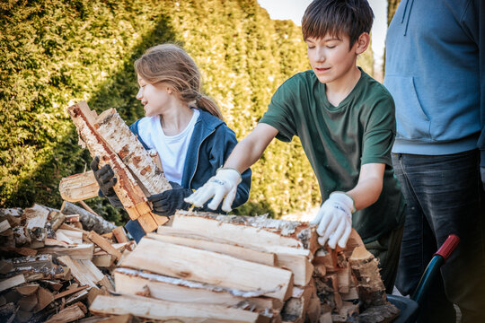 Two children in gloves carry chopped firewood next to a pile of firewood in a sunny courtyard of a residential building, where the house, gate, and parked car are visible.