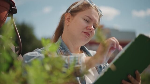 Woman examines plants. Female researcher documents plant details diligently outside. An individual attentively analyzing and recording plant features during sunny outdoor research session