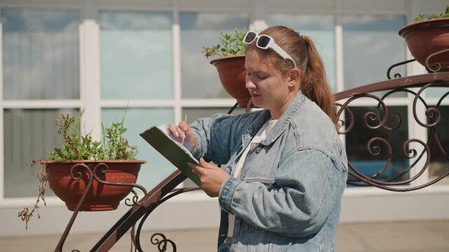 Confident girl reviews her portfolio while standing on balcony surrounded by plants. An assured young woman checks her documents outdoors with potted plants and city windows in background