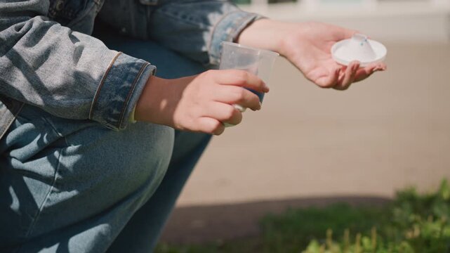 Closeup hands opening white pillbox on curb, denim sleeve and jeans visible, careful fingers checking dosage, natural light, practical selfcare ritual, subtle outdoor setting, focused intimate detail