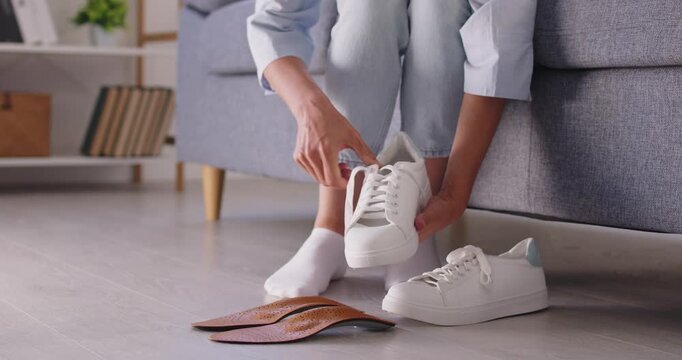 Woman lacing sneakers with orthotic insoles. Sitting at home, she prepares her feet for walking, choosing cushioning support and comfort for posture and pain relief. Soft care for tired feet.