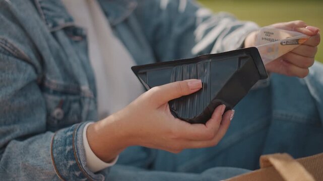Hands holding black takeout container. Denim jacket sleeve on lap, unwrapping meal from plastic lid while seated on grass warm afternoon light, relaxed solo picnic vibe, sustainable packaging and cozy