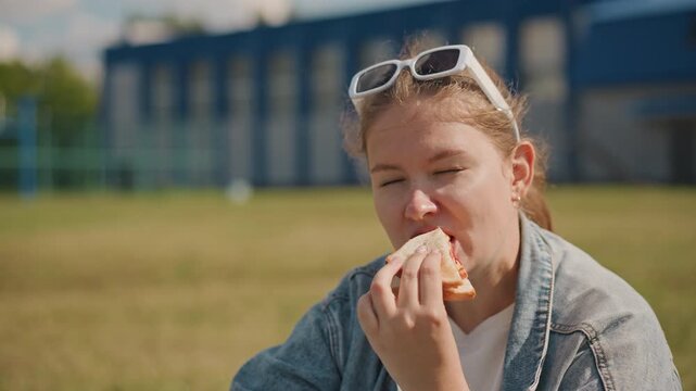 Young woman relaxing with food outdoors. Female student eating lunch on grassy area under bright sky. Woman wearing denim jacket smiling as she bites sandwich on sunny afternoon outdoors
