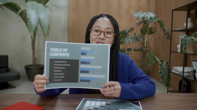 Young Asian female business analyst with braided hair sitting at office desk, holding up printed documents with bar charts and explaining infographics during virtual meeting via video call