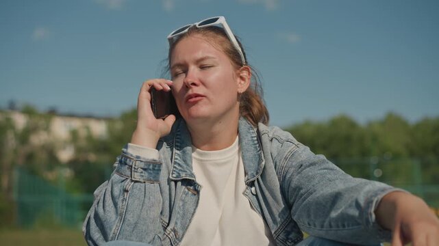 Young woman animated on phone outdoors, denim jacket and sunglasses, expressive hand gestures, sports court fence background, bright blue sky, candid storyteller energy and dynamic narration.