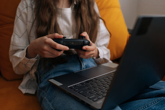 Close-up of young girl hands holding black controller playing video game on laptop. Person in casual clothes sitting on couch at home, enjoying leisure time. Modern hobby and entertainment concept.