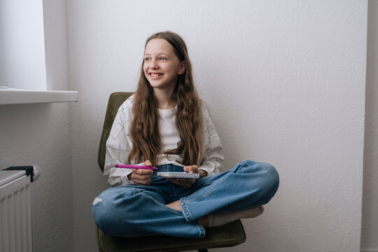 Smiling preteen girl sitting cross legged on chair holding notebook and pen. She looking dreamily out window, feeling happy and inspired. Minimalist interior with natural light. Concept of creativity.
