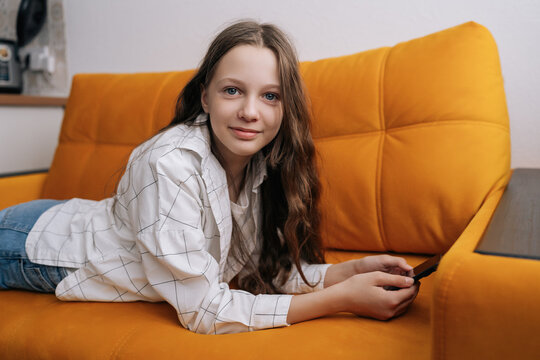 Cute young girl with long brown hair and blue eyes relaxing on bright orange sofa, smiling subtly while holding mobile phone, concept of leisure time and digital connection at home.