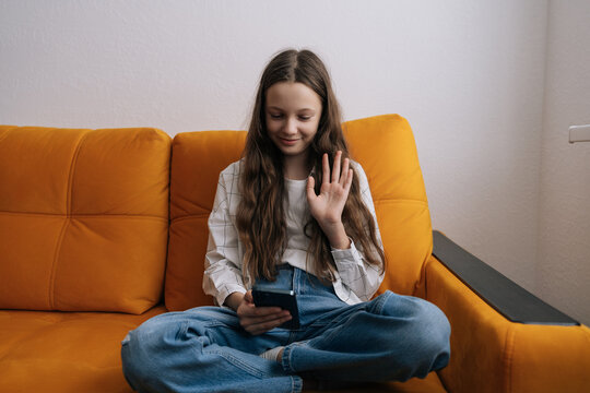 Young preteen girl sitting cross legged on an orange sofa, smiling and waving hand during video call on smartphone, connecting with people remotely from home.