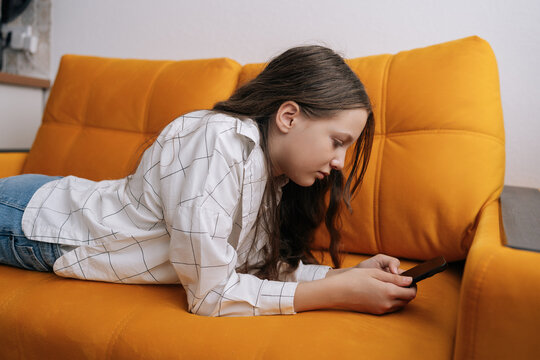Teenager girl lying on yellow couch focusing on smartphone screen, spending leisure time with digital device at home, concept of screen time and technology use among youth.