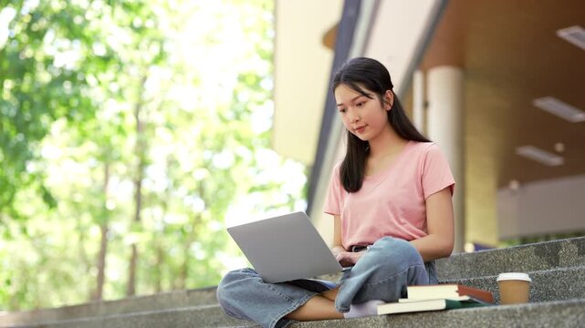 Smiling young asian young woman in university sitting outdoor working on laptop computer working or studying.
