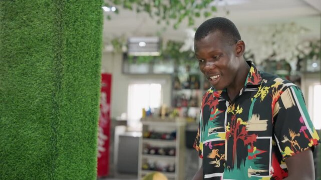 Black man smiling in cafe, casual patterned shirt, greenery wall backdrop, friendly expression, checking phone, waiting for partner, warm modern interior, candid joyful moment, romantic anticipation,