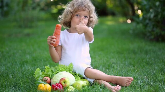 Happy child is sitting outdoors on the grass in summer park. Kid eating carrot against blurred green background. Slow motion