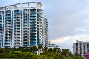 Fototapeta premium Facade of modern white high-rise building with tropical palm trees. Side view of contemporary residential skyscraper architecture with glass balconies in Miami Florida