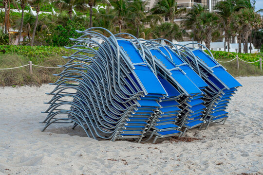 Stacked blue lounge chairs on sandy beach in Miami. Large group of blue metal sunbeds stacked together on the sand with palm trees in the background in Florida.
