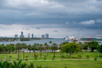 Fototapeta premium Storm clouds over Biscayne Bay and Port of Miami. Wide view of Miami bay with green park in foreground and cruise ships at the port under dramatic dark storm clouds.