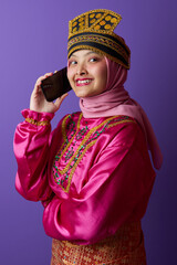 Naklejka premium Smiling Young Woman in Hijab and Traditional Southeast Asian Headdress Talking on Smartphone